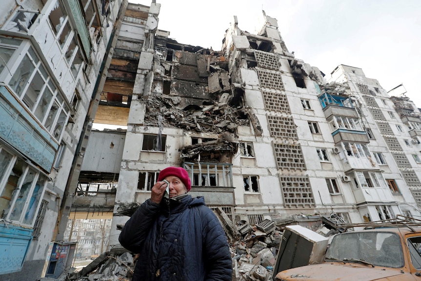 A woman wipes away tears while standing in front of a bombed apartment building. 