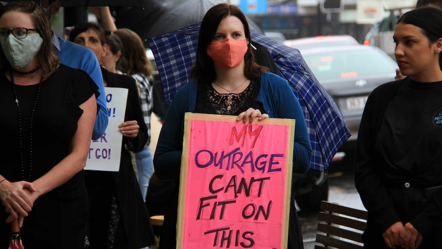 A women wearing a mask holds a sign that reads 'my outrage can't fit on this sign'.