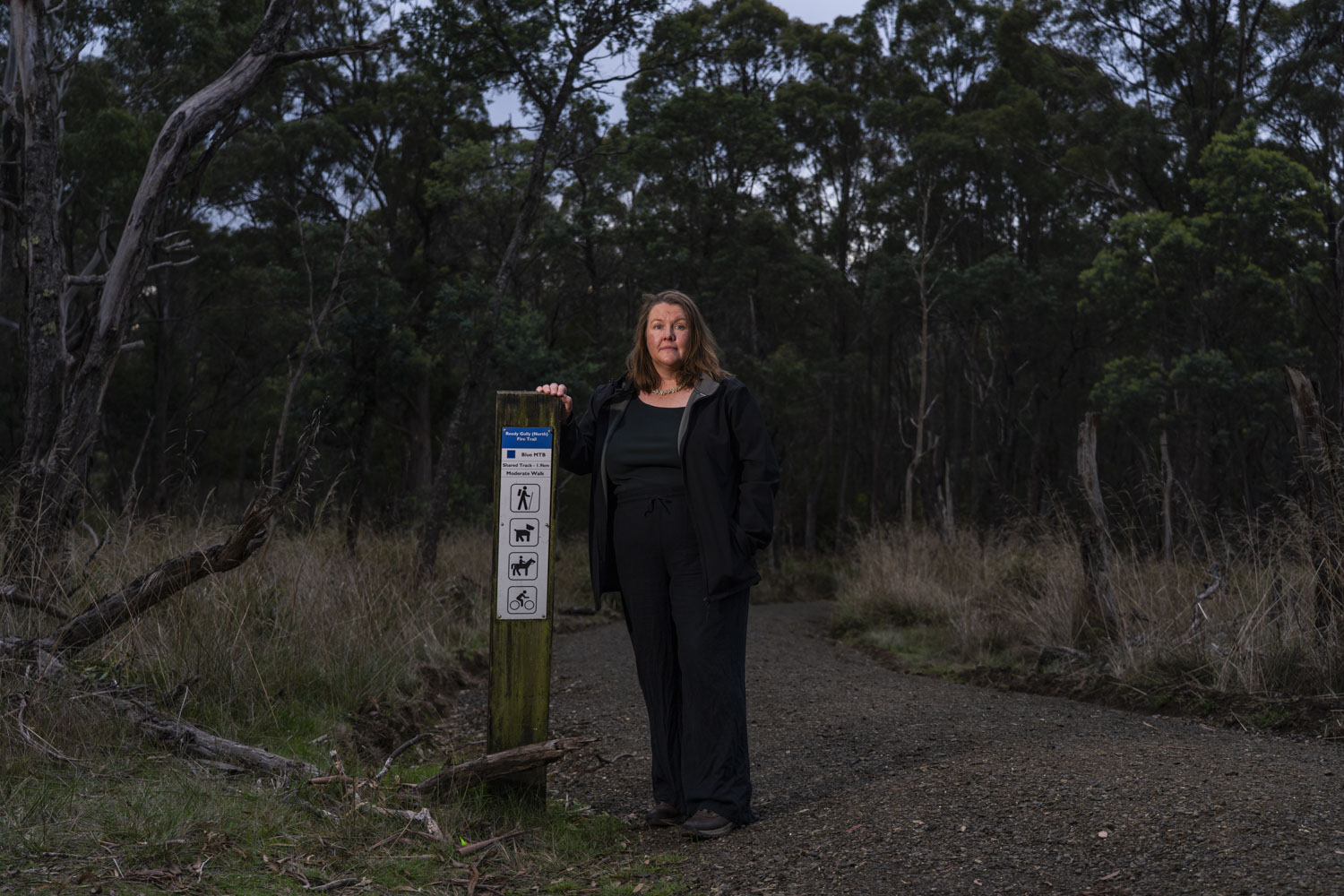a woman with brown hair and a black jacket stands in the bush