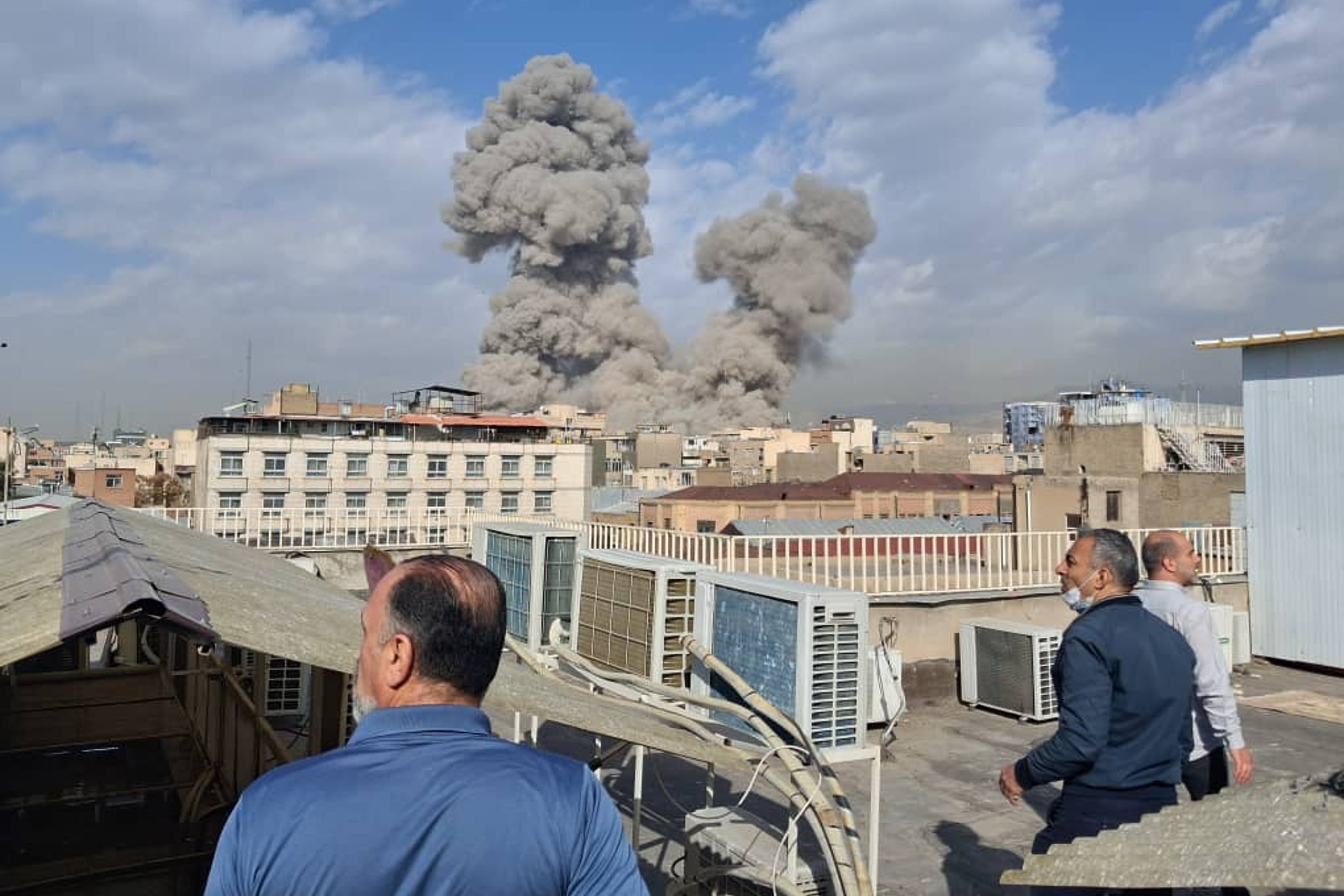Three men watch from a building rooftop as smoke rises on the skyline of a city.