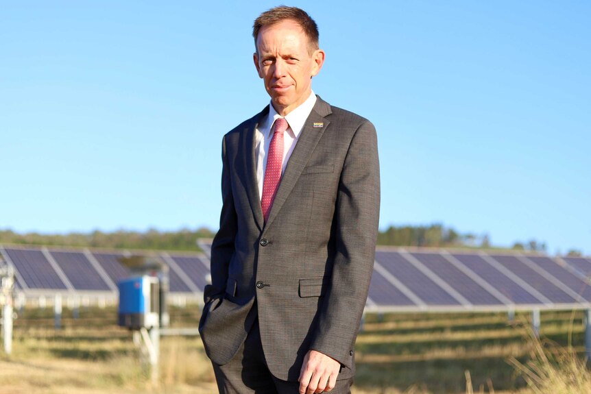 Shane Rattenbury stands smiling slightly in front of some solar panels.