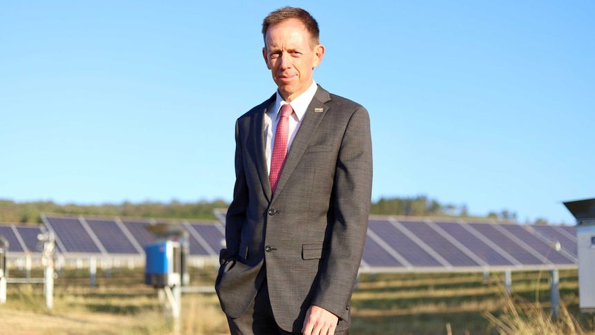 Shane Rattenbury stands smiling slightly in front of some solar panels.