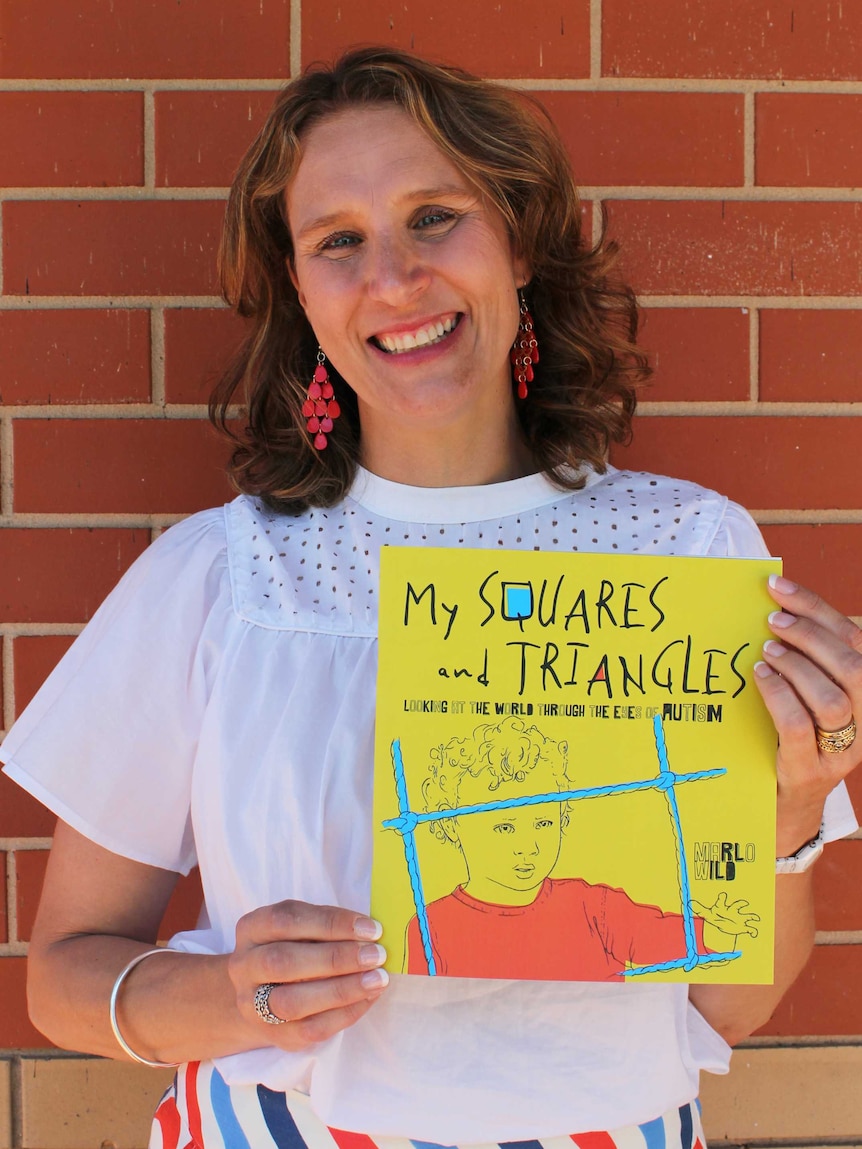 A woman in a white blouse holds a copy of a children's picture book in front of a brick wall.