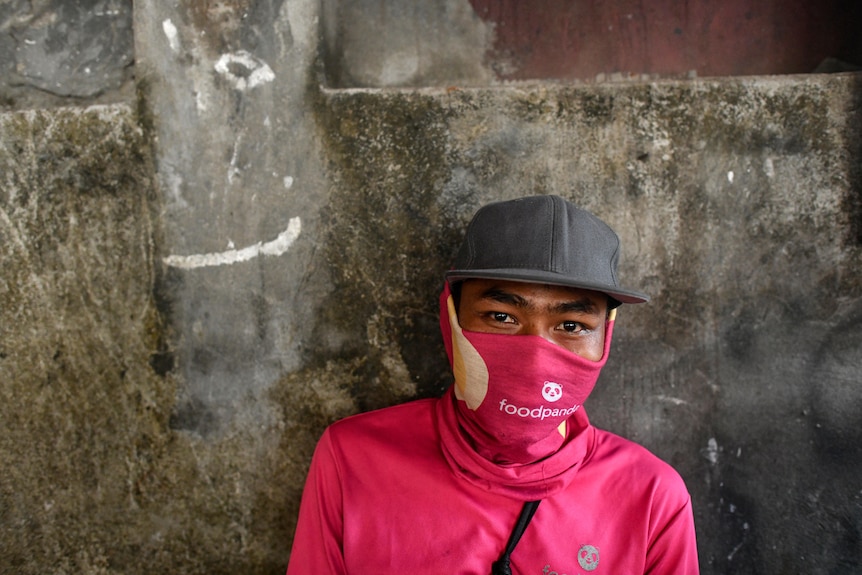 A masked and capped Asian man in pink stands against a stone wall