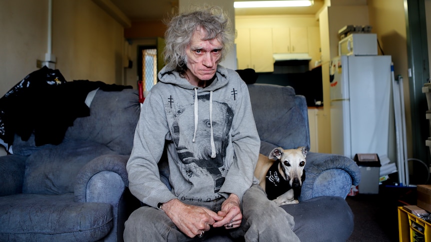 Man with grey jumper and grey hair sits on blue couch next to dog, with apartment kitchen in background