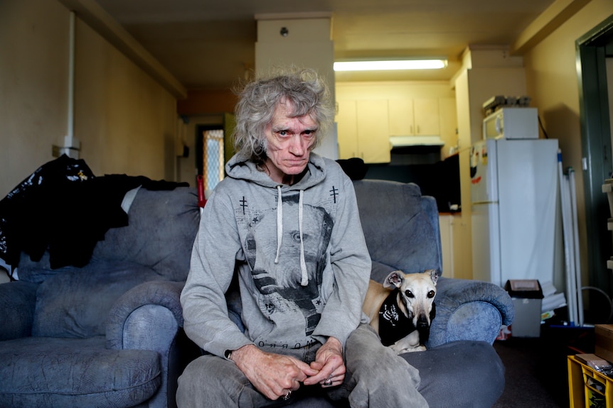 Man with grey jumper and grey hair sits on blue couch next to dog, with apartment kitchen in background