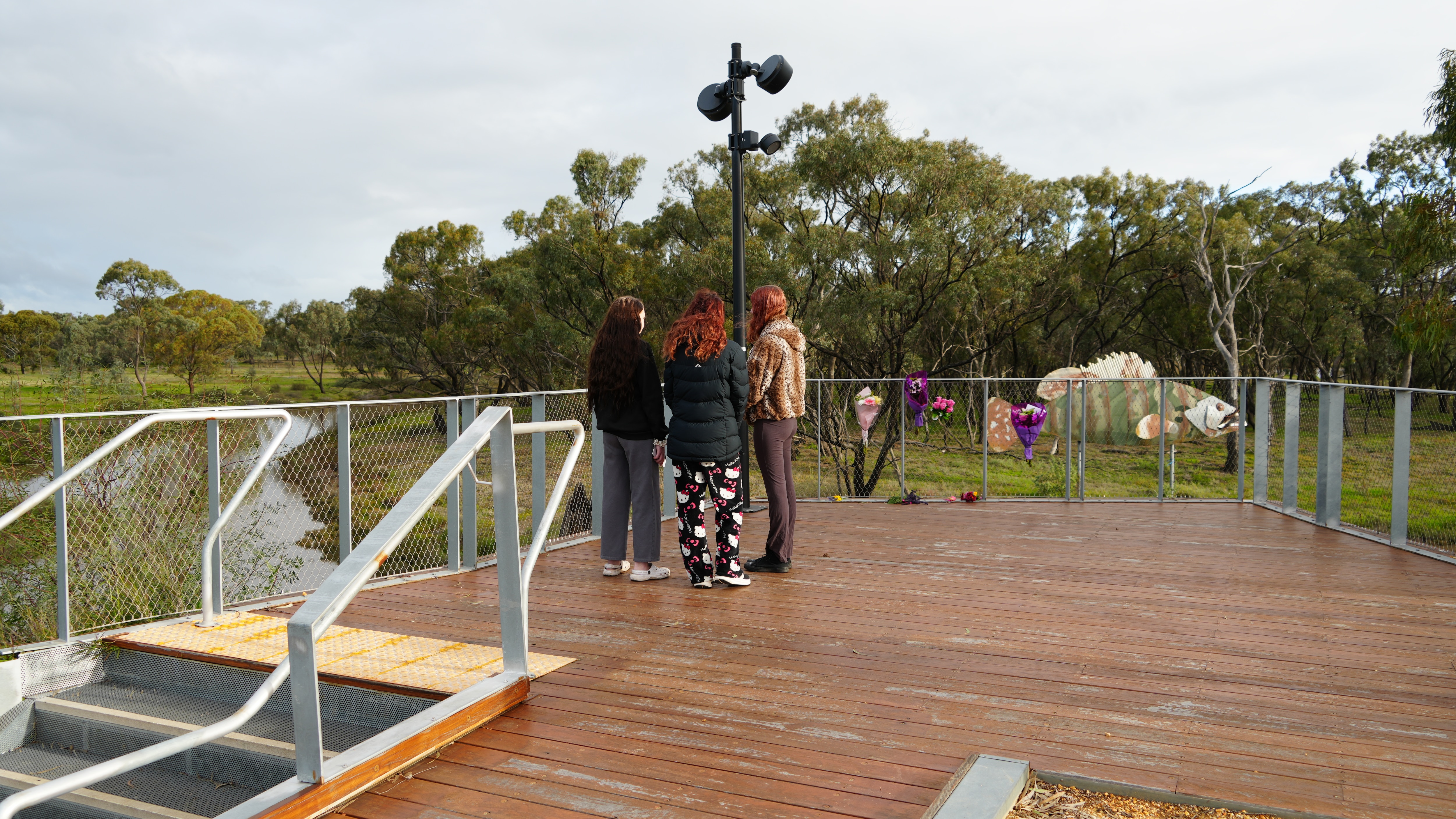 The backs of three young teenage girls wearing tracksuits, metres from flowers at riverside lookout.