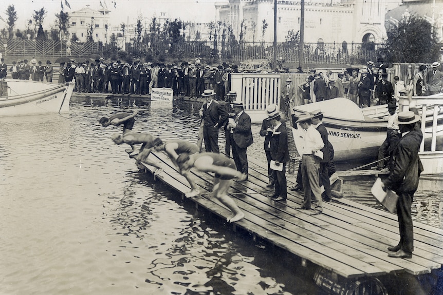 Four men prepare to dive from a wooden pontoon on a lake, around them are men wearing early 20th Century clothes and hats.