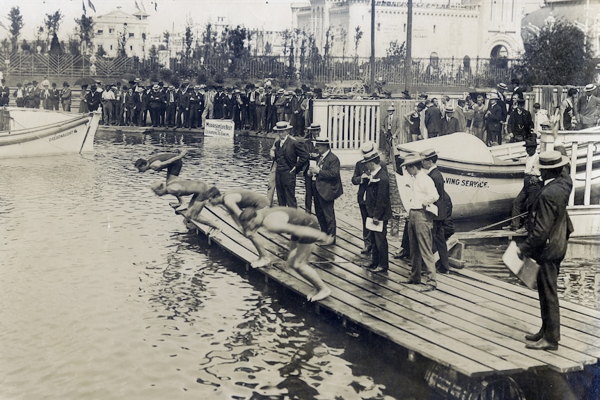 Cuatro hombres se preparan para zambullirse desde un pontón de madera en un lago, y alrededor de ellos hay hombres con ropa y sombreros de principios del siglo XX.