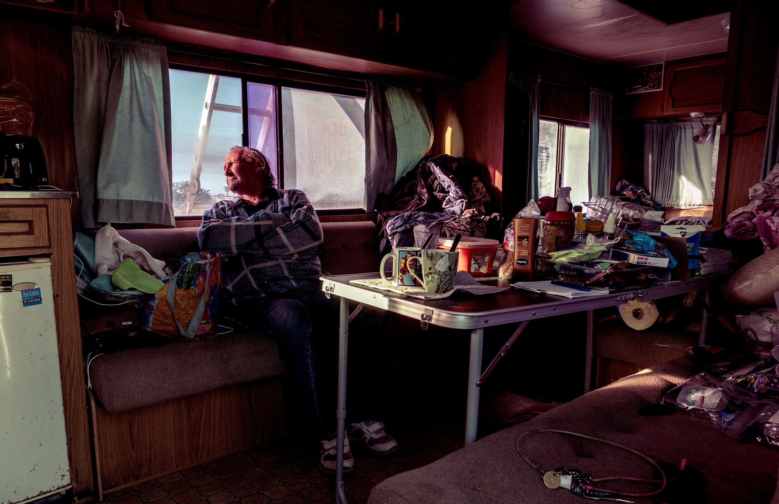 A man sits inside a cluttered caravan, looking out the window at the sunny sky.
