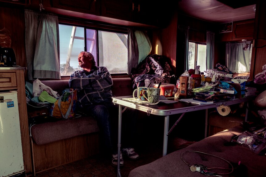 A man sits inside a cluttered caravan, looking out the window at the sunny sky.