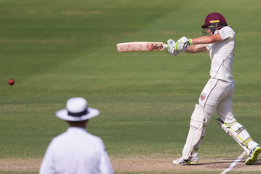 Marnus Labuschagne stretches his bat out at shoulder height in a swing at the ball. He is wearing a red Queensland Bulls helmet.