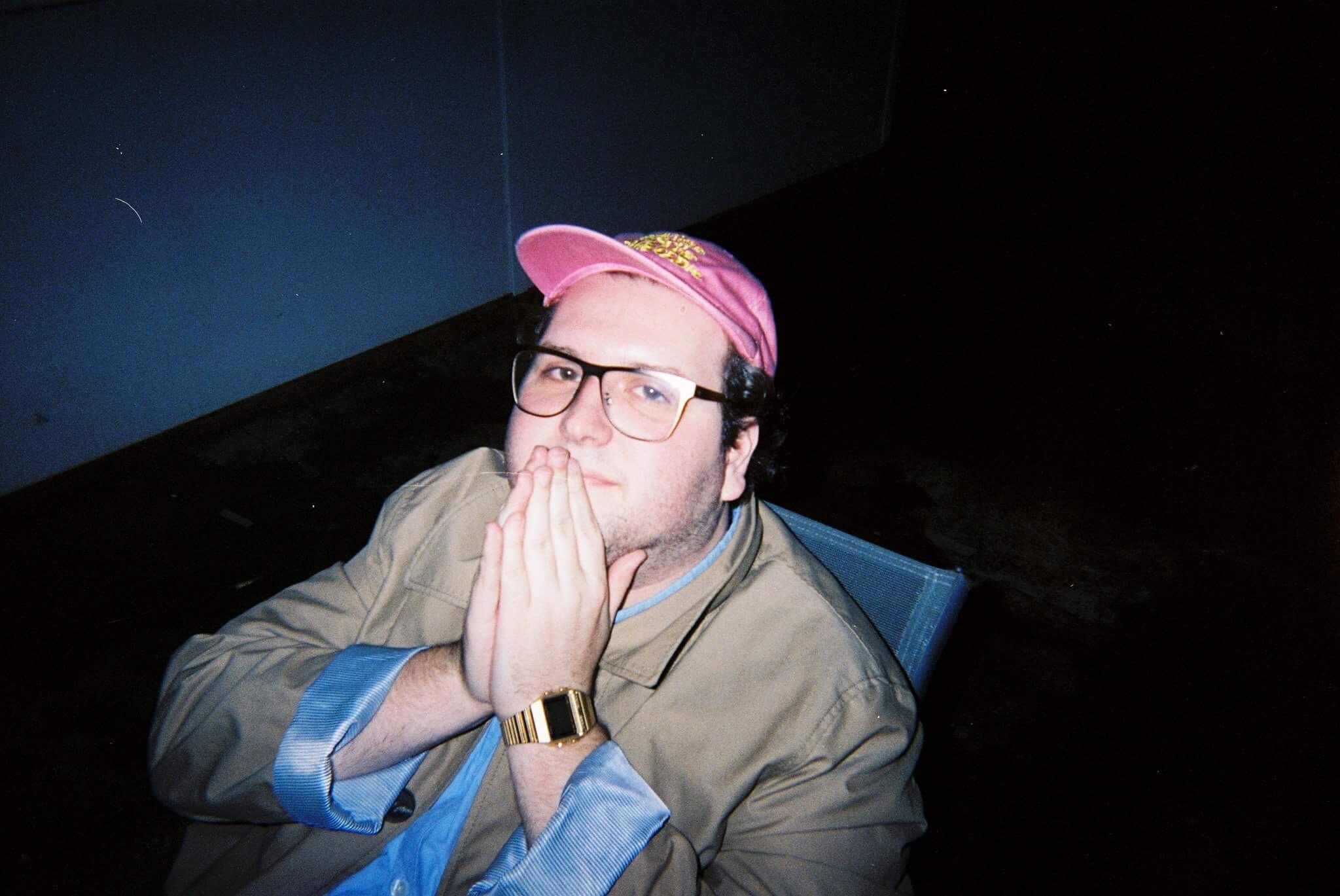 Alexei sits in a chair against a black background, the camera above him, putting his hands to his face in a praying motion.