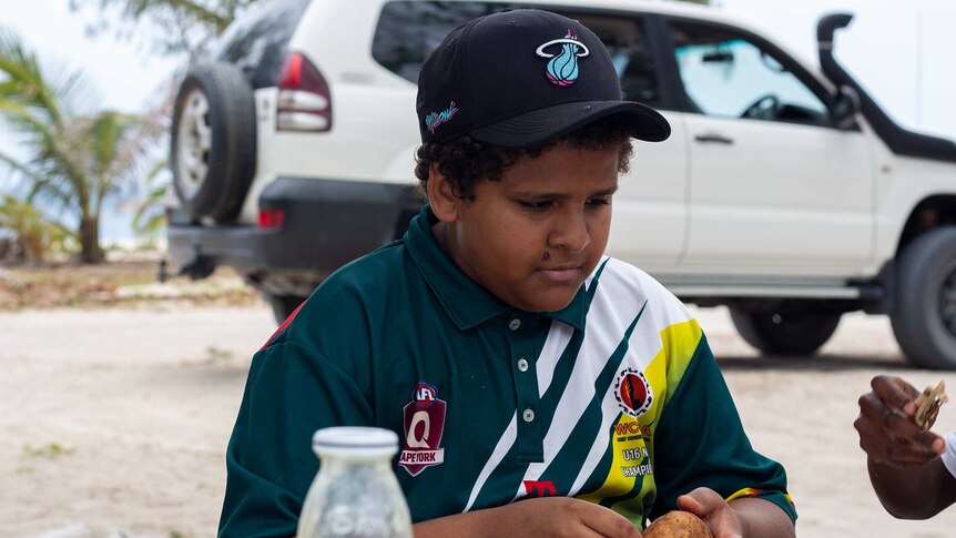 A young boy in a cap sitting outside in front of a large car.