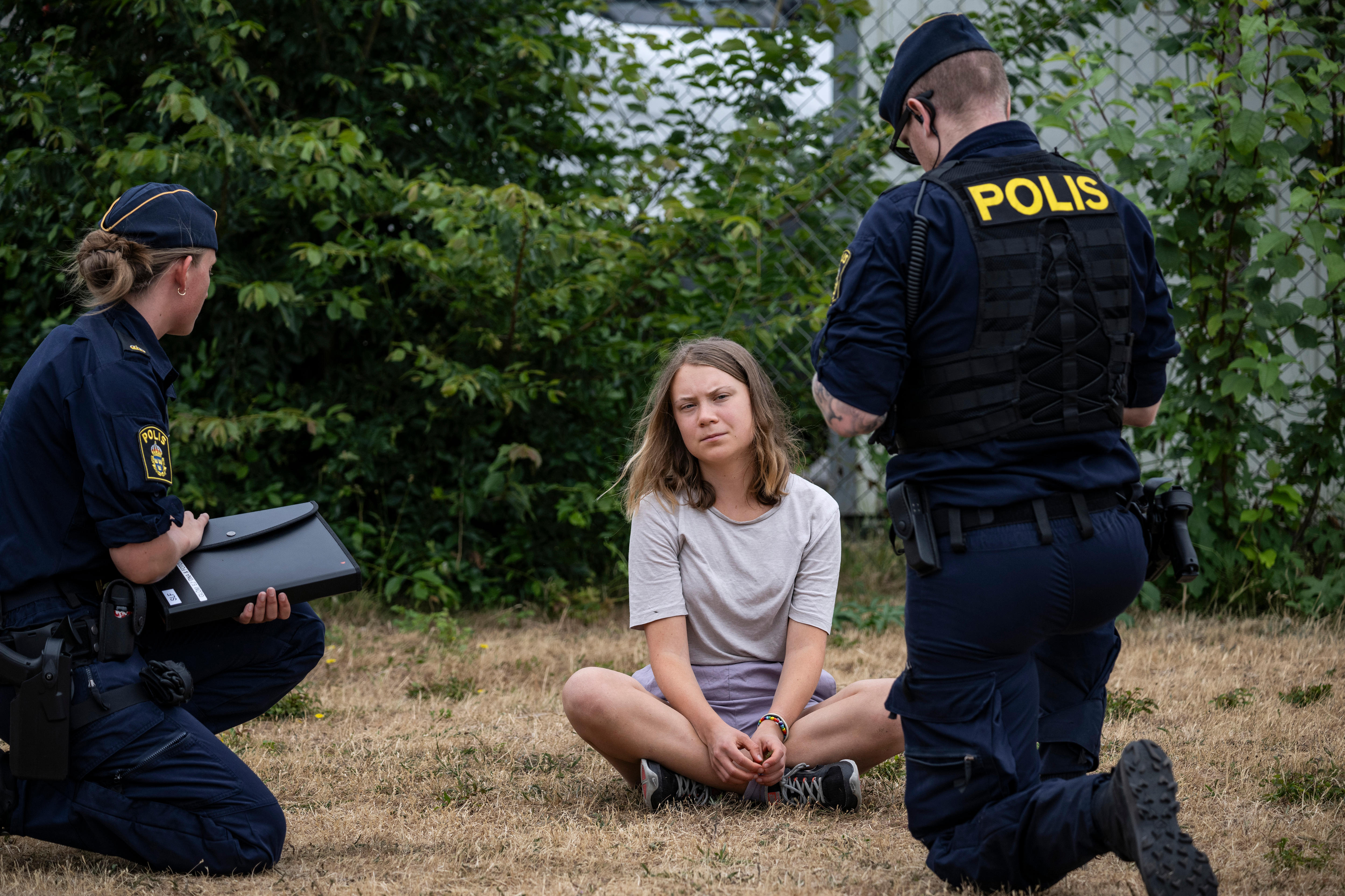 A young woman dressed in T-shirt, shorts and sneakers sits cross-legged on dry grass and speaks to two police officers.