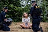 A young woman dressed in T-shirt, shorts and sneakers sits cross-legged on dry grass and speaks to two police officers.