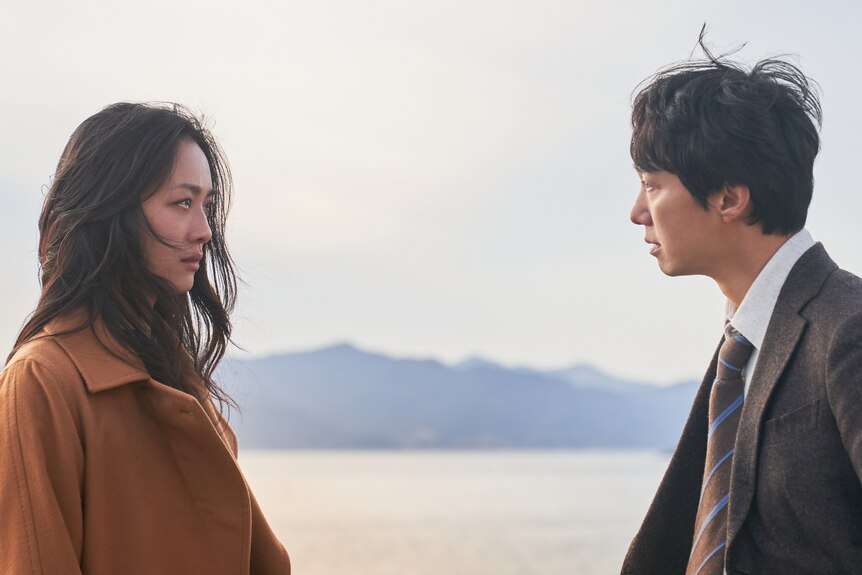 East Asian man and woman facing each other in dramatic stare with the sea and mountains in the background.