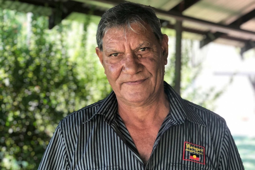 An Indigenous man stands on a veranda with trees behind him, looking at the camera. 