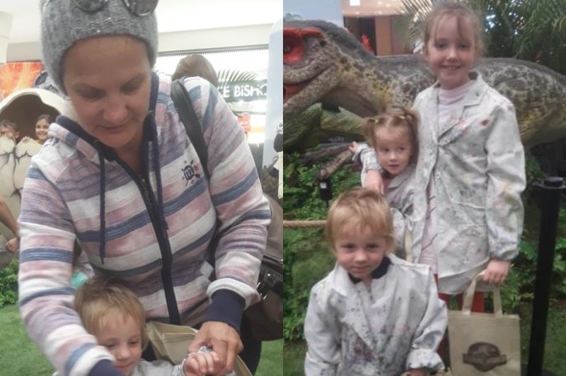 A woman holds the hands of a little boy and three children pose together in front of a dinosaur statue