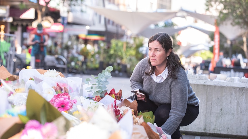 a woman crouches to drop off flowers at a makeshift memorial in Bondi Junction outside of the shopping centre