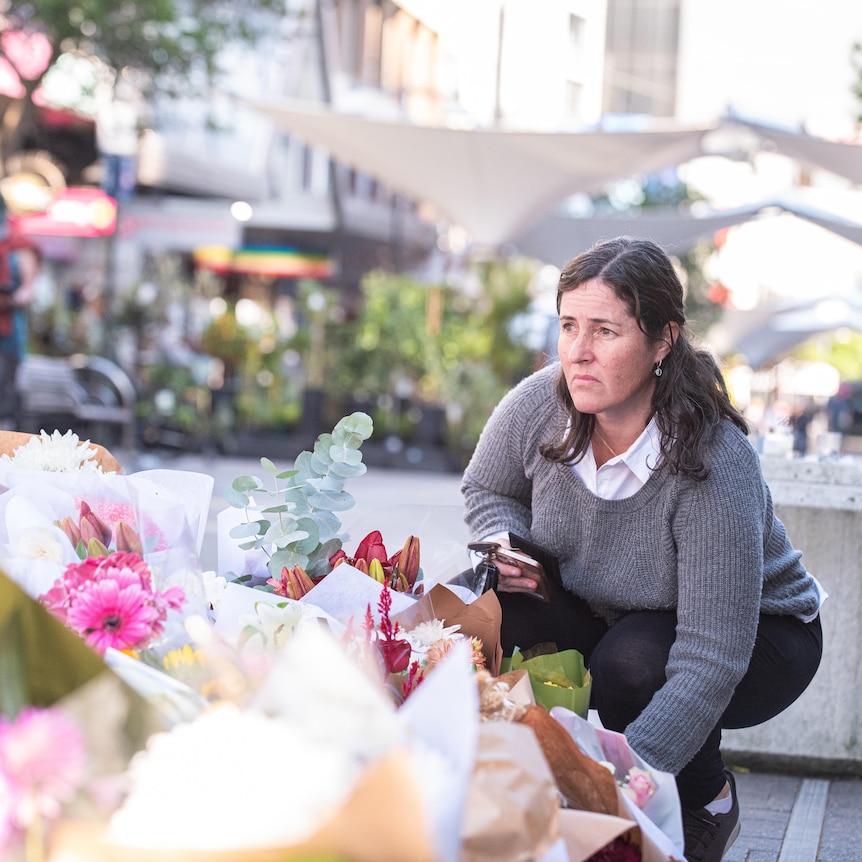 a woman crouches to drop off flowers at a makeshift memorial in Bondi Junction outside of the shopping centre