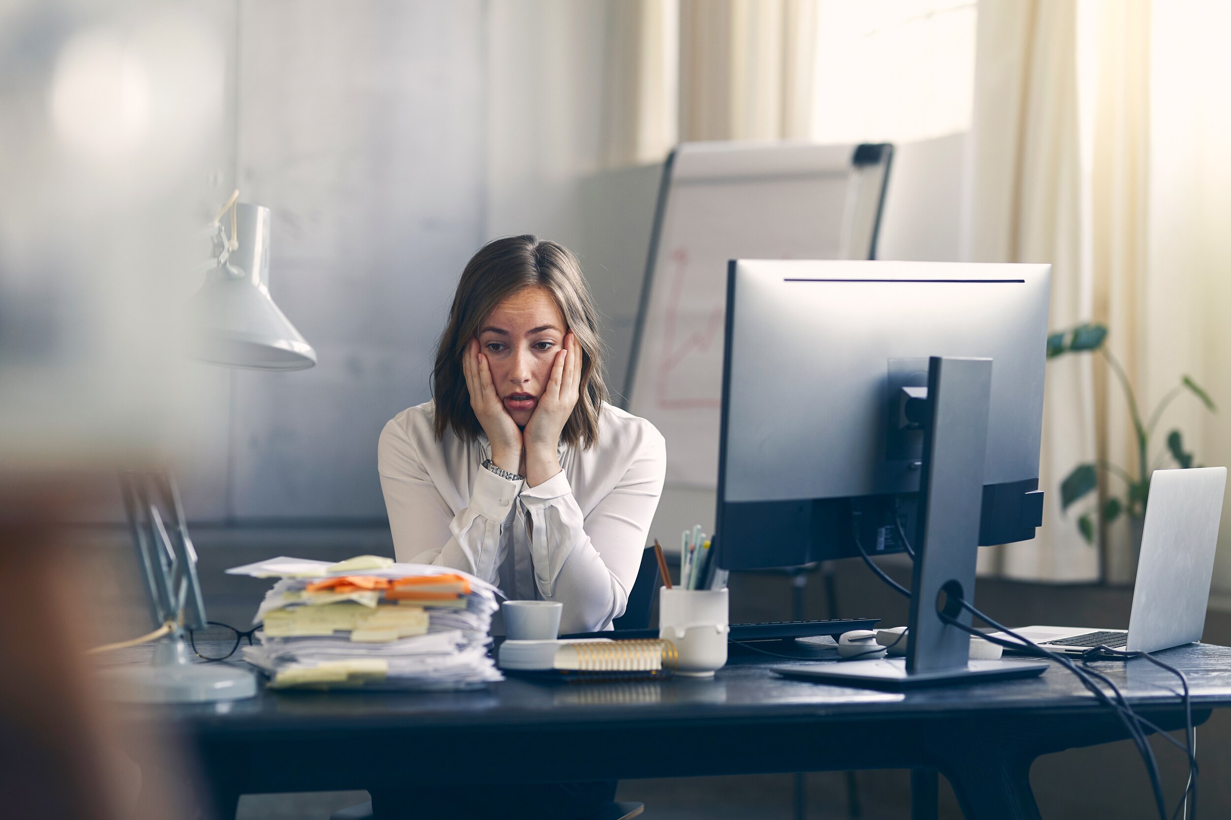 Brunette white woman at her desk looking very stressed in a corporate office job looking at a stack of papers