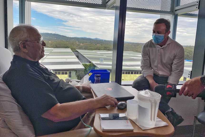 Richard sits up in a hospital bed talking to the health minister.