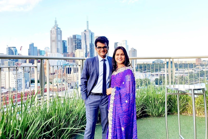 A smiling woman in purple saree stands close to a man in a blue suit on a balcony, with plants, Melbourne skyline in background.