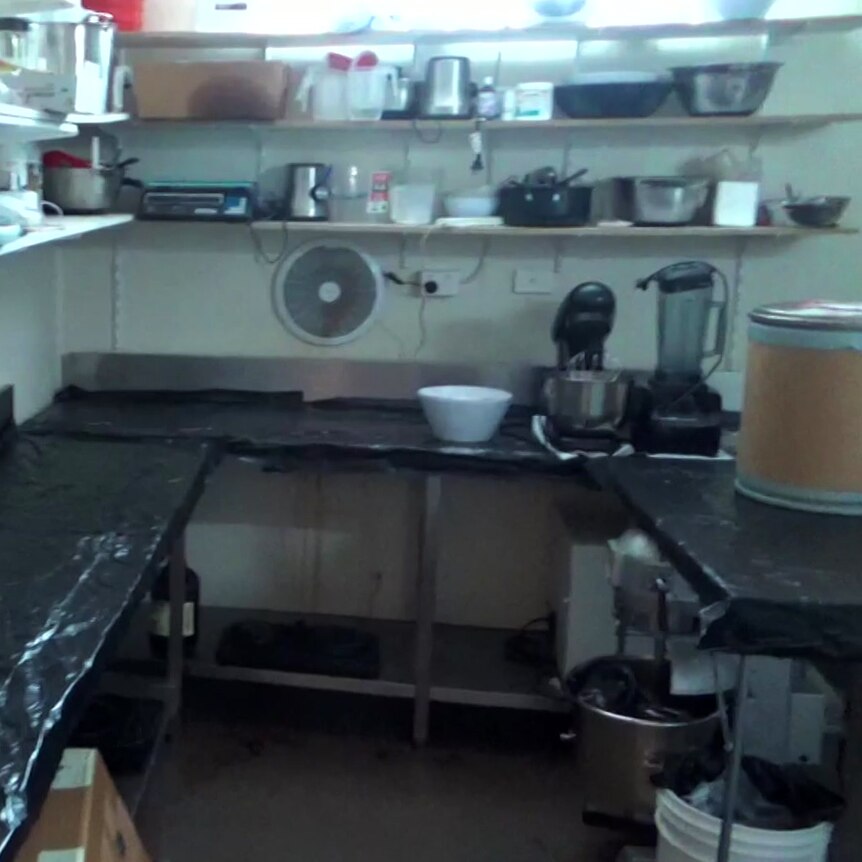  Cluttered shelves line a wall above a bench lined with black plastic. A blender and kitchen mixer are on the bench.