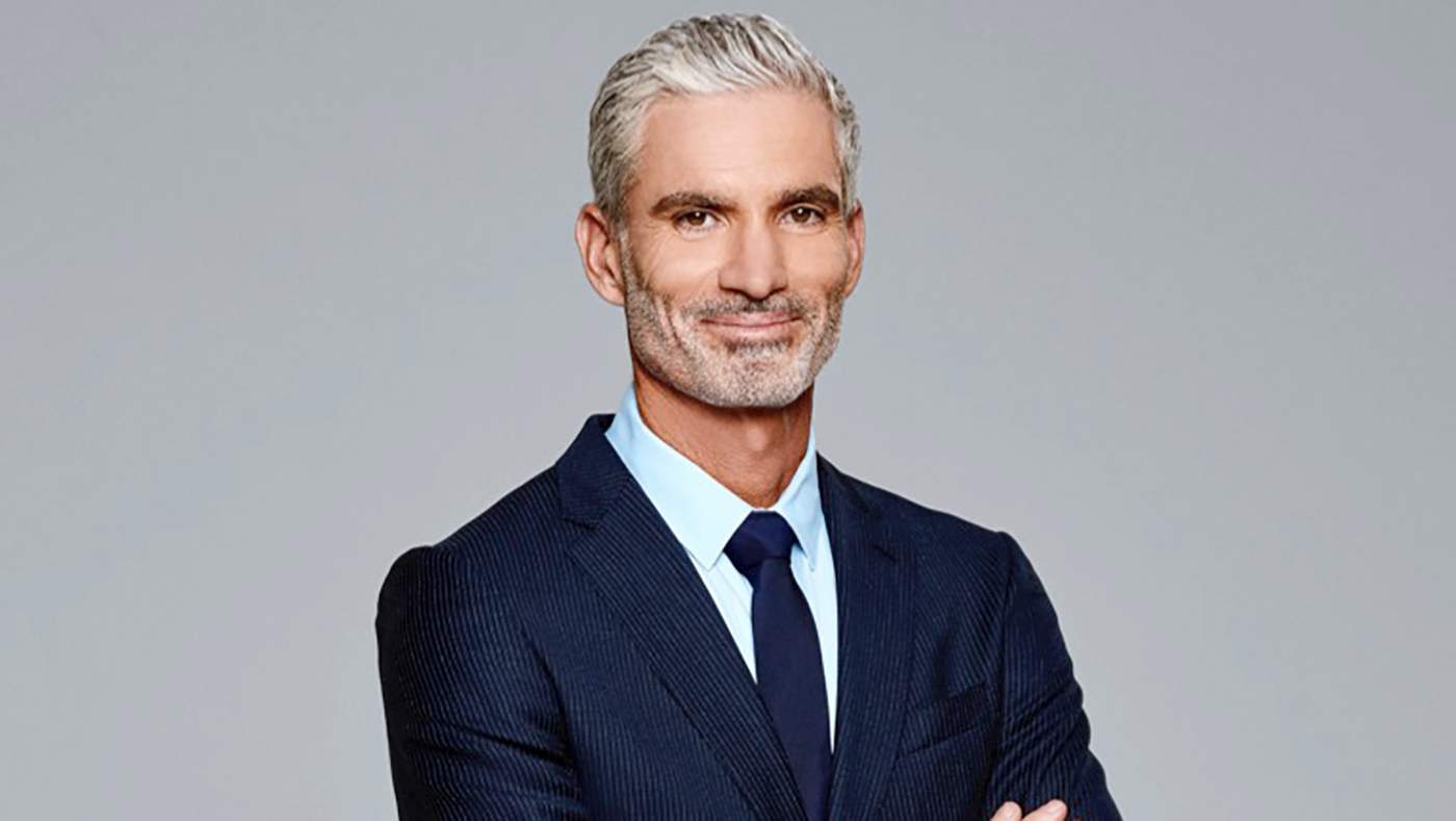 Craig Foster wearing a blue suit with his arms crossed and smiling in front of a gray background