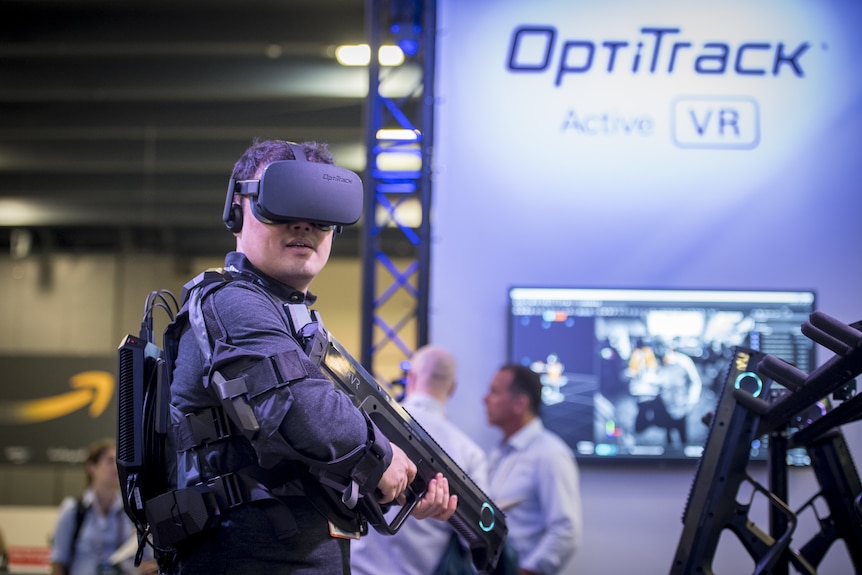 A man wears a virtual reality headset and holds a toy gun as he plays a video game at a conference.