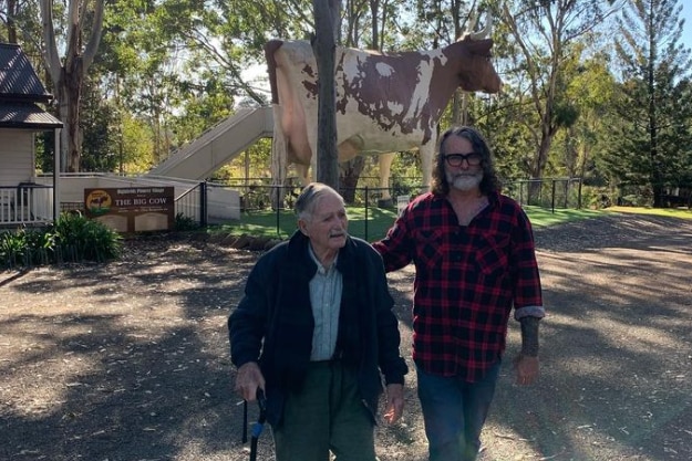 Older man and elderley man walk with big cow in background