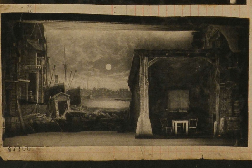 A black and white photo of a painted backdrop that shows run down houses on a dock alongside a shack with a table and two chairs
