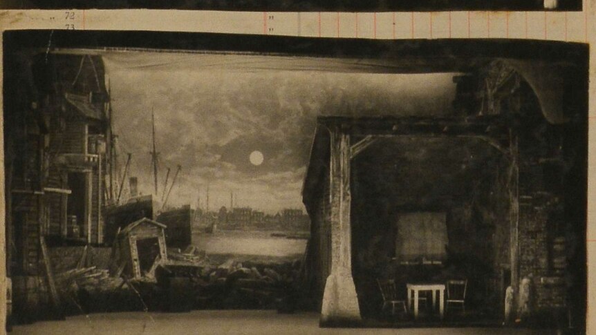 A black and white photo of a painted backdrop that shows run down houses on a dock alongside a shack with a table and two chairs