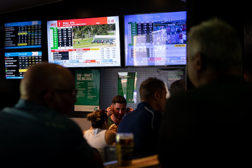 A wide shot of men sitting at a dark bar with large TV screens showing horse-racing in the background.