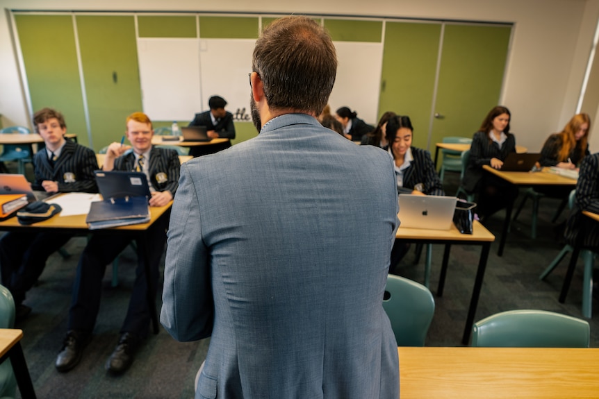 A teacher in a grey suit jacket is talking to a class of about 10 students.