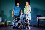 Actors Michael Kazonis and Evelyn Tsavalas posing with a wheelchair on stage.