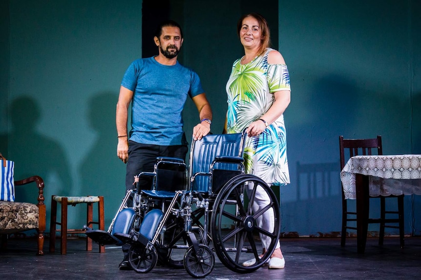 Actors Michael Kazonis and Evelyn Tsavalas posing with a wheelchair on stage.