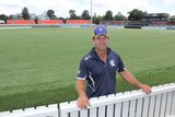 Man stands behind white cricket fence in front of Manuka oval