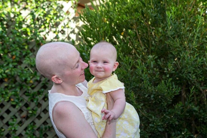 Catherine smiles while holding her daughter. She has no hair on her head as a result of cancer treatment.