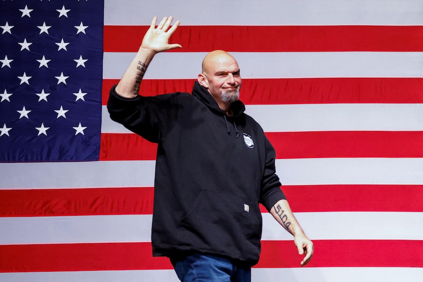 A bald bearded man in a hoodie waves while walking past a US flag backdrop