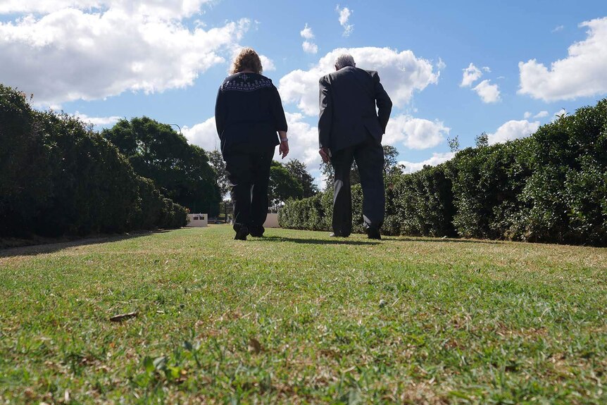 Two people walk through a grassy clearing bound by shrubs at Ipswich cemetery.