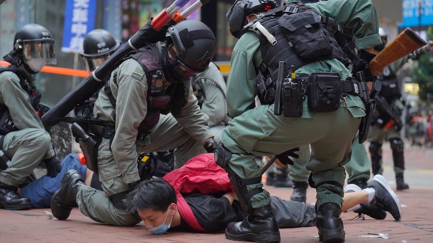 police in riot gear hold down a protester in a red shirt