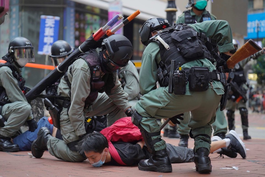 police in riot gear hold down a protester in a red shirt