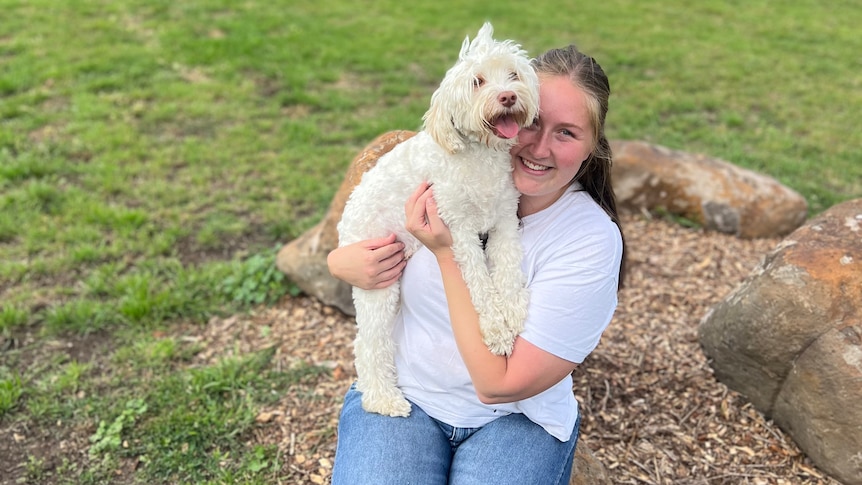 Woman in white shirt and blue jeans hugs a white labradoodle in a grassy field. They are both smiling.