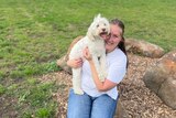 Woman in white shirt and blue jeans hugs a white labradoodle in a grassy field. They are both smiling.