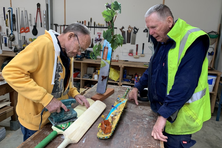 Brett Wiltshire stands at workbench and levels out an old cricket bat with Rutherglen Men's Shed President Ern Walder looking on