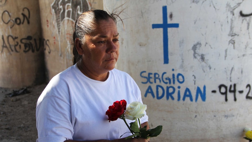 The mother of Sergio Hernandez holds flowers standing at the spot her son was shot dead where his name