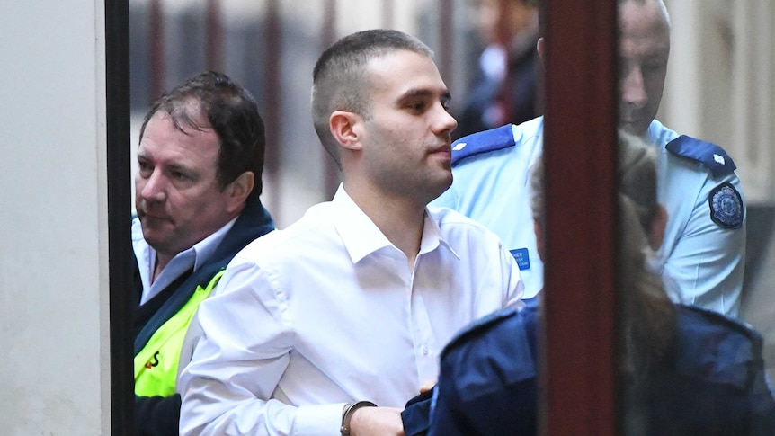 A man with short brown hair, in a white collared shirt, is escorted into a building by prison guards.