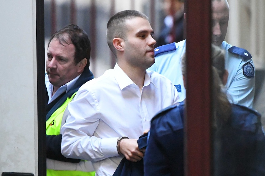 A man with short brown hair, in a white collared shirt, is escorted into a building by prison guards.